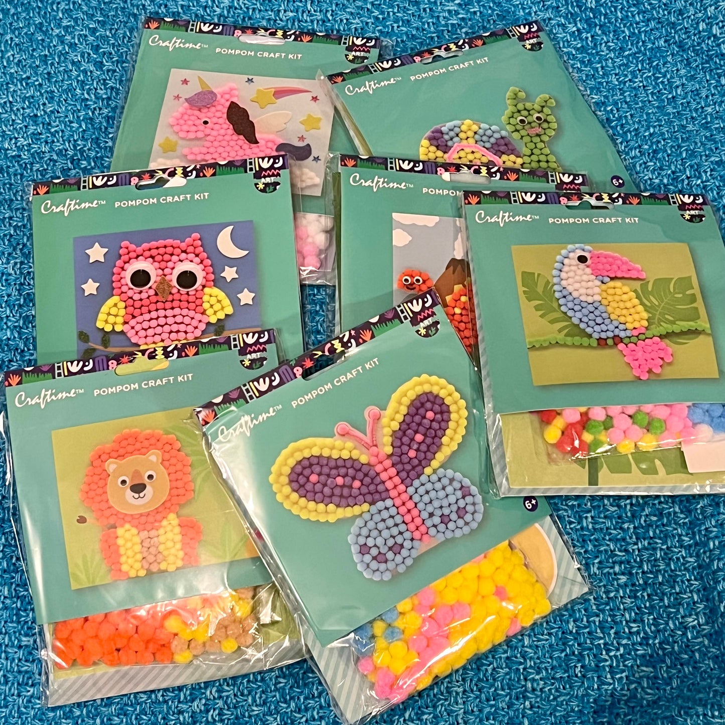 Collection of Craftime pom-pom craft kits on a blue textured background